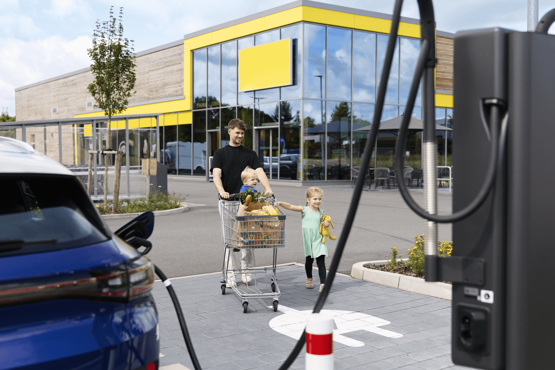 Father and children in front of supermarket. Charging car in the front. 