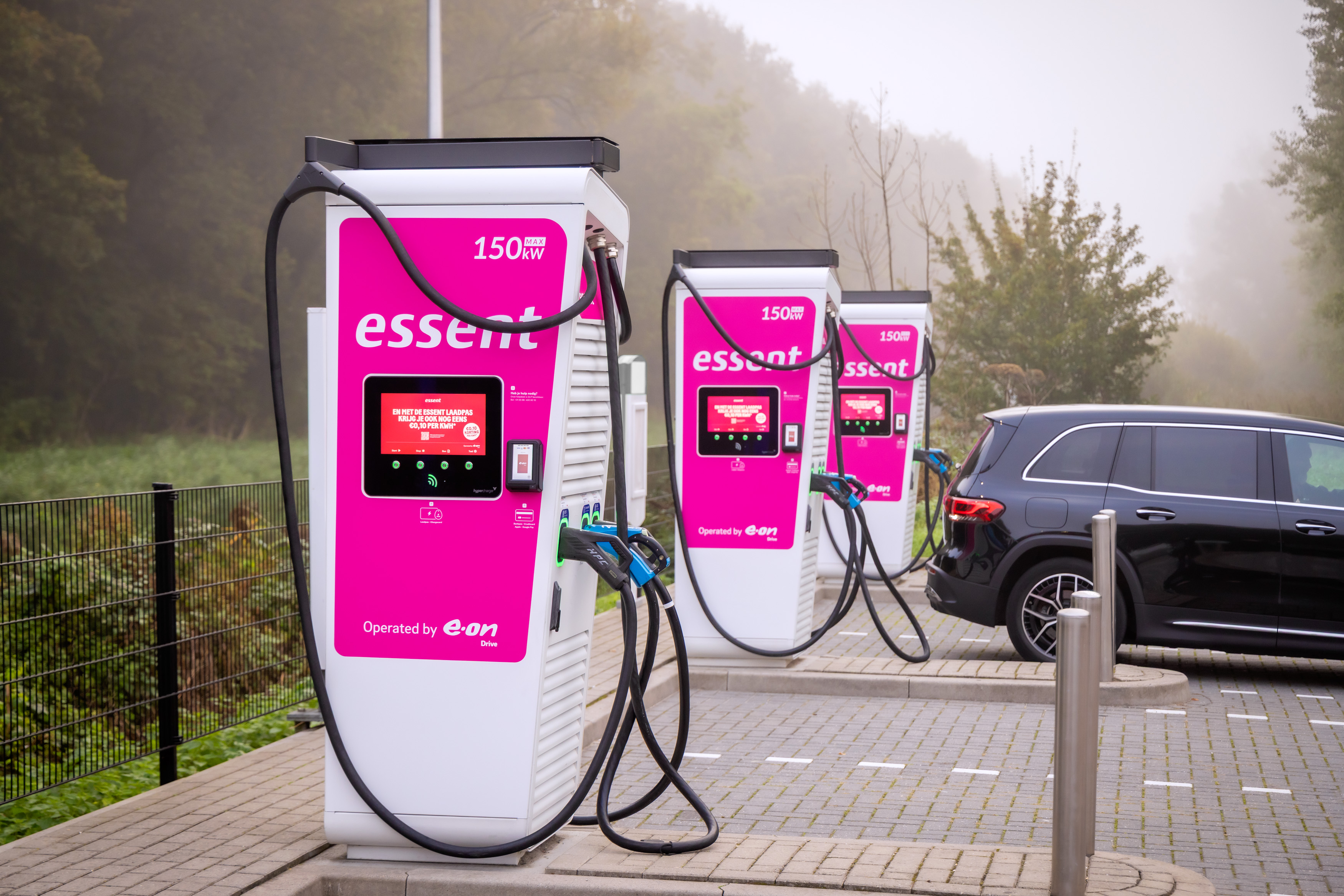 Three Essent 150 kW fast chargers marked "Operated by E.ON Drive," with a black SUV parked at the far charger on a foggy day.